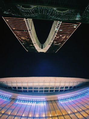 A futuristic architectural structure with illuminated patterns. The design features symmetrical lines and vibrant lighting that create a captivating, modern aesthetic. The reflection enhances the visual impact, lending a sense of depth and symmetry to the scene.