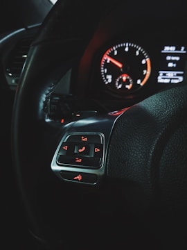 A close-up of a car's steering wheel with control buttons on it, including phone call and volume symbols. The dashboard is visible in the background, showing a speedometer with a red needle and a digital display with various readings.