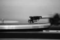 A close-up of a vinyl record player focusing on the needle and tonearm poised above a rotating record. The image captures the texture of the grooves and the sleek design of the turntable, rendered in black and white.