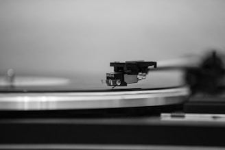 A close-up of a vinyl record player focusing on the needle and tonearm poised above a rotating record. The image captures the texture of the grooves and the sleek design of the turntable, rendered in black and white.
