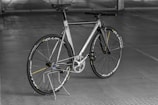A sleek, modern bicycle with a metallic frame and thin tires, set against an indoor concrete backdrop. The design is minimalist with a focus on speed and performance, highlighted by aerodynamic features and a lightweight structure. The bicycle stands upright on a simple metal stand.