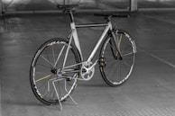 A sleek, modern bicycle with a metallic frame and thin tires, set against an indoor concrete backdrop. The design is minimalist with a focus on speed and performance, highlighted by aerodynamic features and a lightweight structure. The bicycle stands upright on a simple metal stand.