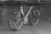 A sleek, modern bicycle with a metallic frame and thin tires, set against an indoor concrete backdrop. The design is minimalist with a focus on speed and performance, highlighted by aerodynamic features and a lightweight structure. The bicycle stands upright on a simple metal stand.