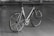 A sleek, modern bicycle with a metallic frame and thin tires, set against an indoor concrete backdrop. The design is minimalist with a focus on speed and performance, highlighted by aerodynamic features and a lightweight structure. The bicycle stands upright on a simple metal stand.
