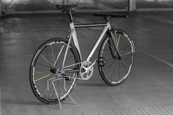 A sleek, modern bicycle with a metallic frame and thin tires, set against an indoor concrete backdrop. The design is minimalist with a focus on speed and performance, highlighted by aerodynamic features and a lightweight structure. The bicycle stands upright on a simple metal stand.