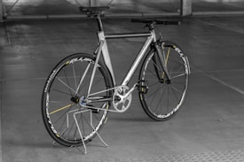 A sleek, modern bicycle with a metallic frame and thin tires, set against an indoor concrete backdrop. The design is minimalist with a focus on speed and performance, highlighted by aerodynamic features and a lightweight structure. The bicycle stands upright on a simple metal stand.