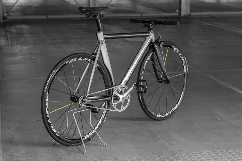 A sleek, modern bicycle with a metallic frame and thin tires, set against an indoor concrete backdrop. The design is minimalist with a focus on speed and performance, highlighted by aerodynamic features and a lightweight structure. The bicycle stands upright on a simple metal stand.
