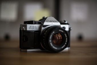 A vintage Canon AE-1 film camera with a large lens is prominently displayed. It rests on a wooden surface, with a blurred background contributing to a shallow depth of field.