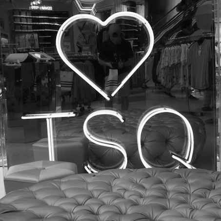 A black and white photo featuring a shop's interior with several clothing racks in the background. In the foreground, there is tufted seating with a neon sign saying 'I ❤️ TSQ'. A person wearing a cap is reflected in the glass front of the shop, holding a shopping bag.