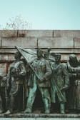 A bronze statue features a group of determined-looking individuals in historical attire. The central figure holds a flag, symbolizing leadership or a call to action. The statue is situated against a stone wall, with small branches protruding from the top, adding a touch of nature.