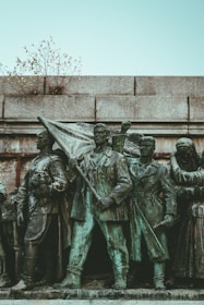A bronze statue features a group of determined-looking individuals in historical attire. The central figure holds a flag, symbolizing leadership or a call to action. The statue is situated against a stone wall, with small branches protruding from the top, adding a touch of nature.
