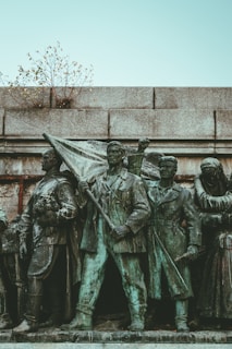 A bronze statue features a group of determined-looking individuals in historical attire. The central figure holds a flag, symbolizing leadership or a call to action. The statue is situated against a stone wall, with small branches protruding from the top, adding a touch of nature.
