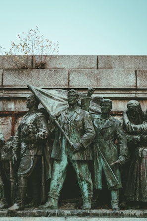 A bronze statue features a group of determined-looking individuals in historical attire. The central figure holds a flag, symbolizing leadership or a call to action. The statue is situated against a stone wall, with small branches protruding from the top, adding a touch of nature.