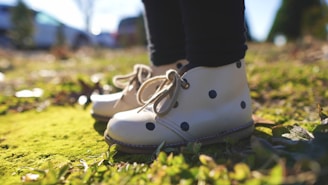 A pair of cream-colored boots with black polka dots on a child's feet. The boots have laces and are positioned on a vibrant green mossy ground, with the background out of focus indicating an outdoor setting.