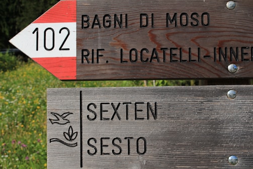 A wooden signpost with two sections, each pointing in different directions. The top sign has a red and white triangular pattern with '102' and text in Italian referring to locations like 'Bagni di Moso' and 'Rif. Locatelli-Innerkofler.' The bottom sign reads 'Sexten Sesto' in black lettering and features a floral emblem. The background has green foliage and wildflowers.