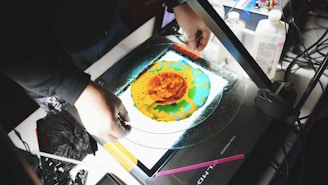 A person is holding a circular glass plate with vibrant colors, including orange, yellow, and green, spread in a fluid design on its surface. The setup appears to be on a lightbox or projector with various equipment and materials around, including bottles, cables, and a black plastic bag.