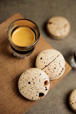A glass filled with espresso is placed on a wooden surface, accompanied by speckled, cream-colored macarons. The macarons have dark spots, adding a contrasting visual texture. One macaron is broken, revealing its interior. The background is out of focus, allowing the coffee and macarons to stand out.