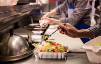 A kitchen setting where food preparation is underway. A person in a checkered shirt and blue apron is adding fresh vegetables to a foil container using a spoon. The counter is lined with other containers and kitchen tools, creating a busy and organized atmosphere.