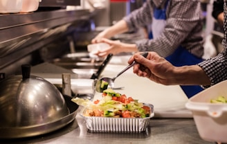 A kitchen setting where food preparation is underway. A person in a checkered shirt and blue apron is adding fresh vegetables to a foil container using a spoon. The counter is lined with other containers and kitchen tools, creating a busy and organized atmosphere.
