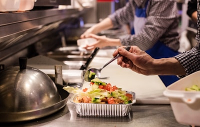 A kitchen setting where food preparation is underway. A person in a checkered shirt and blue apron is adding fresh vegetables to a foil container using a spoon. The counter is lined with other containers and kitchen tools, creating a busy and organized atmosphere.