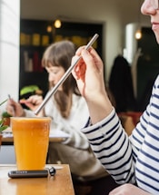 A person wearing a striped shirt is using a metal straw to drink from a glass of orange juice. Another person is in the background, focused on their task, with blurred bookshelves behind them. The setting appears to be a cozy cafe with warm lighting.