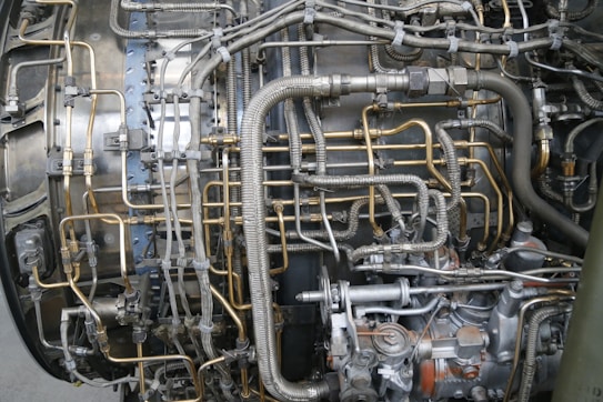 An intricate network of metallic pipes and tubes is interconnected around a complex mechanical structure, featuring a combination of silver, gray, and brass-colored elements. The setup suggests a section of an industrial machine or a jet engine, with numerous valves, joints, and connectors.