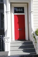 A bright red door is set within a white architectural frame on an exterior of a building. The number 844 is displayed above the door. The entrance is accessed by a short set of gray stone steps. The surrounding walls are painted light gray and feature classical detailing.
