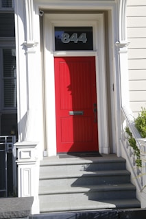 A bright red door is set within a white architectural frame on an exterior of a building. The number 844 is displayed above the door. The entrance is accessed by a short set of gray stone steps. The surrounding walls are painted light gray and feature classical detailing.
