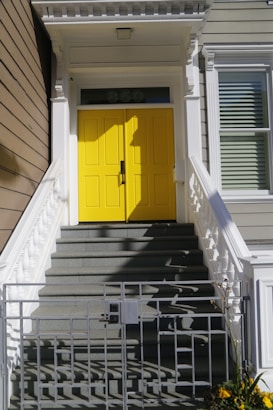 Bright yellow double doors are centrally located at the top of a set of gray, shadowed stairs. These stairs are bordered by white railings on both sides. A small metal gate is visible at the bottom, slightly open. The number 850 is displayed above the doors in a transom window. The surrounding walls are a muted beige color with classic, white trim and molding details.