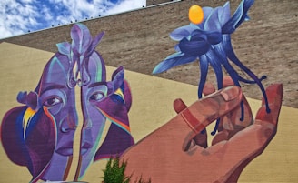 A large, colorful mural painted on a brick wall depicts an abstract representation of a human face, composed of blue, purple, and orange tones. A hand holds a surreal blue flower with a bright yellow center, extending towards the right. The background consists of earthy brown and beige hues, adding contrast to the vivid colors in the artwork.