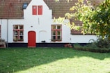 A charming house with a white facade and a red door and window frames. The roof is made of brown tiles, and there are several potted plants placed near the windows. The foreground features a lush, green lawn with some trees partially shading the area.