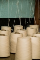 Multiple spools of white thread are arranged in rows, with thin threads extending upwards. The background consists of a wall with a blue and black section, and part of a brown wooden object is visible on the right side.
