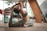 A person is practicing yoga indoors, performing a backbend on a wooden floor. The individual is wearing a matching set of athletic wear, and the room contains various pieces of furniture and decor, including shelves with items, a potted plant, and a stack of folded yoga mats.