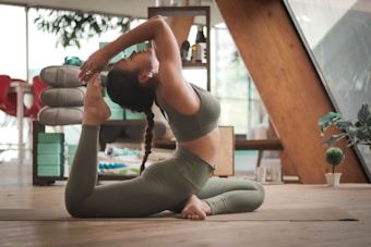 A person is practicing yoga indoors, performing a backbend on a wooden floor. The individual is wearing a matching set of athletic wear, and the room contains various pieces of furniture and decor, including shelves with items, a potted plant, and a stack of folded yoga mats.