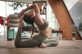 A person is practicing yoga indoors, performing a backbend on a wooden floor. The individual is wearing a matching set of athletic wear, and the room contains various pieces of furniture and decor, including shelves with items, a potted plant, and a stack of folded yoga mats.