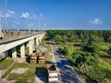 A large highway overpass is seen on the left side, elevated with multiple supporting pillars. Below the overpass, several trucks are parked on a patch of grassy land. To the right, a narrow road is bordered by lush green trees and vegetation, extending into the distance under a bright blue sky with scattered clouds.