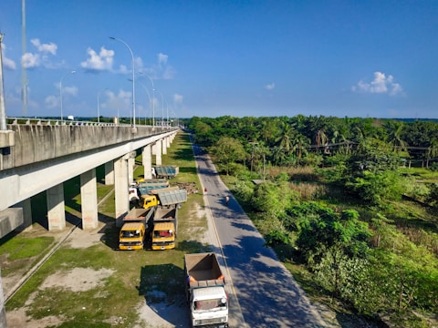 A large highway overpass is seen on the left side, elevated with multiple supporting pillars. Below the overpass, several trucks are parked on a patch of grassy land. To the right, a narrow road is bordered by lush green trees and vegetation, extending into the distance under a bright blue sky with scattered clouds.