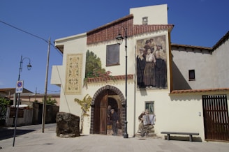A beige building features two large, detailed murals on its walls. To the left, a mural depicts a vine with leaves climbing beside a rustic wooden door. Above, a mural showcases a group of people in traditional clothing, with expressions ranging from content to serious. An accordion player is painted sitting near a bench on the wall. The scene is set in an empty street with street lamps, a bench, and a parking sign.