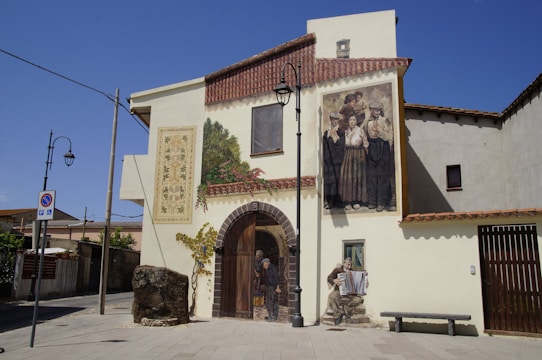 A beige building features two large, detailed murals on its walls. To the left, a mural depicts a vine with leaves climbing beside a rustic wooden door. Above, a mural showcases a group of people in traditional clothing, with expressions ranging from content to serious. An accordion player is painted sitting near a bench on the wall. The scene is set in an empty street with street lamps, a bench, and a parking sign.