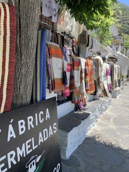 Several colorful woven blankets hang on a white stone wall along a rustic street. The blankets feature stripes and patterns in hues of red, orange, blue, purple, and brown. A tree trunk stands to the left, and overhanging green foliage provides partial shade. A sign with partial text 'FÁBRICA DE MERMELADAS' is visible in the foreground, hinting at a local market or shop setting. Clothing items hang further down the wall.