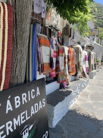 Several colorful woven blankets hang on a white stone wall along a rustic street. The blankets feature stripes and patterns in hues of red, orange, blue, purple, and brown. A tree trunk stands to the left, and overhanging green foliage provides partial shade. A sign with partial text 'FÁBRICA DE MERMELADAS' is visible in the foreground, hinting at a local market or shop setting. Clothing items hang further down the wall.