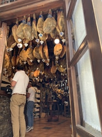 Hanging cured meats are seen in a shop, with legs of ham suspended from the ceiling. Two men stand at the counter, one in a white shirt and khaki pants. The interior displays a rustic ambiance, with products lining the shelves and barrels adding to the decor.