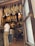 Hanging cured meats are seen in a shop, with legs of ham suspended from the ceiling. Two men stand at the counter, one in a white shirt and khaki pants. The interior displays a rustic ambiance, with products lining the shelves and barrels adding to the decor.