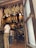 Hanging cured meats are seen in a shop, with legs of ham suspended from the ceiling. Two men stand at the counter, one in a white shirt and khaki pants. The interior displays a rustic ambiance, with products lining the shelves and barrels adding to the decor.