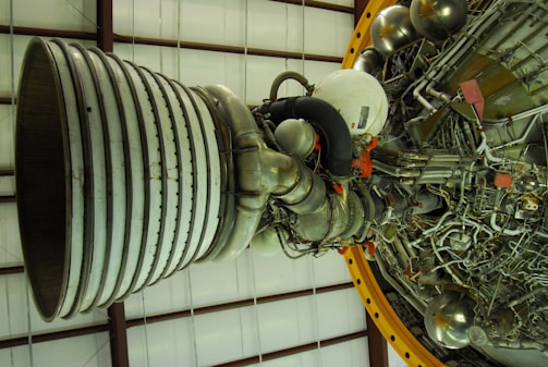 A large and complex rocket engine displays intricate arrays of pipes, cables, and metallic structures. The engine bell is prominent, featuring gray and metallic tones, with various components interconnected. The background consists of a grid-like structure, possibly a part of a large facility or hangar.