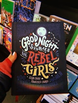 A hand holds a book titled 'Good Night Stories for Rebel Girls' surrounded by other colorful books on a display. The cover features bold, whimsical typography and illustrations including a moon, stars, and constellation motifs.