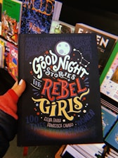 A hand holds a book titled 'Good Night Stories for Rebel Girls' surrounded by other colorful books on a display. The cover features bold, whimsical typography and illustrations including a moon, stars, and constellation motifs.