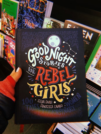 A hand holds a book titled 'Good Night Stories for Rebel Girls' surrounded by other colorful books on a display. The cover features bold, whimsical typography and illustrations including a moon, stars, and constellation motifs.