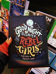 A hand holds a book titled 'Good Night Stories for Rebel Girls' surrounded by other colorful books on a display. The cover features bold, whimsical typography and illustrations including a moon, stars, and constellation motifs.