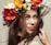 A woman is adorned with a vibrant floral crown featuring red, white, yellow, and orange flowers. Her long brown hair flows naturally around her face as she gazes softly at the viewer with a subtle smile. The background is a neutral color, highlighting the colorful flowers and her expressive features.